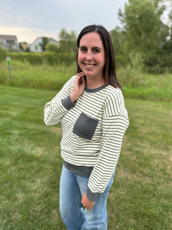 A woman standing outdoors on a grassy lawn wearing a cream and dark gray striped long-sleeve top with a solid dark gray chest pocket and matching dark gray cuffs and hem, paired with blue jeans.