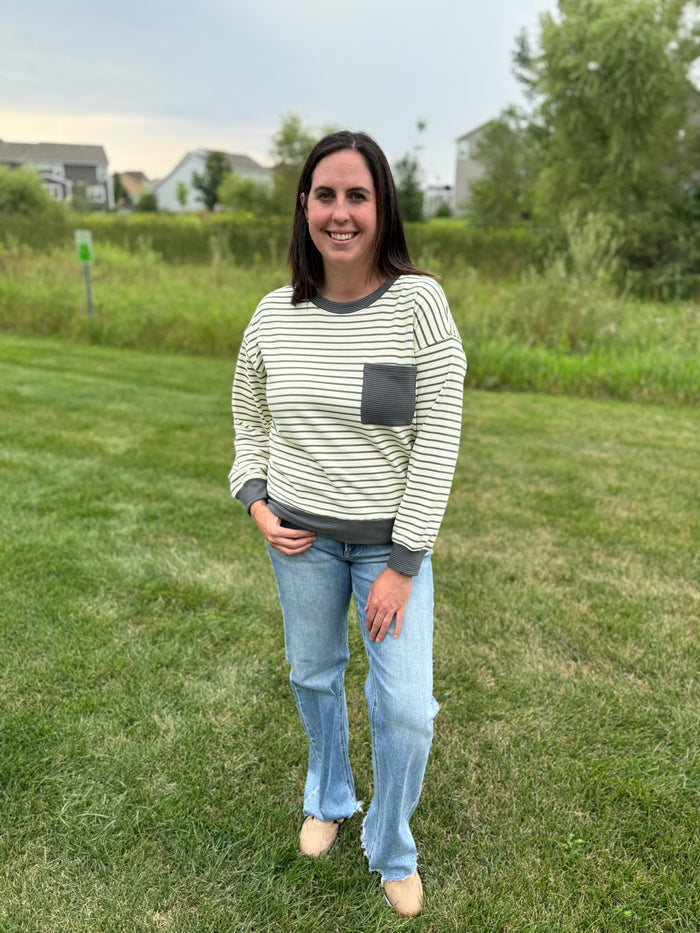 A woman standing on a grassy lawn wearing a cream and dark green striped long-sleeve top with a dark gray pocket, paired with light blue jeans and tan shoes.