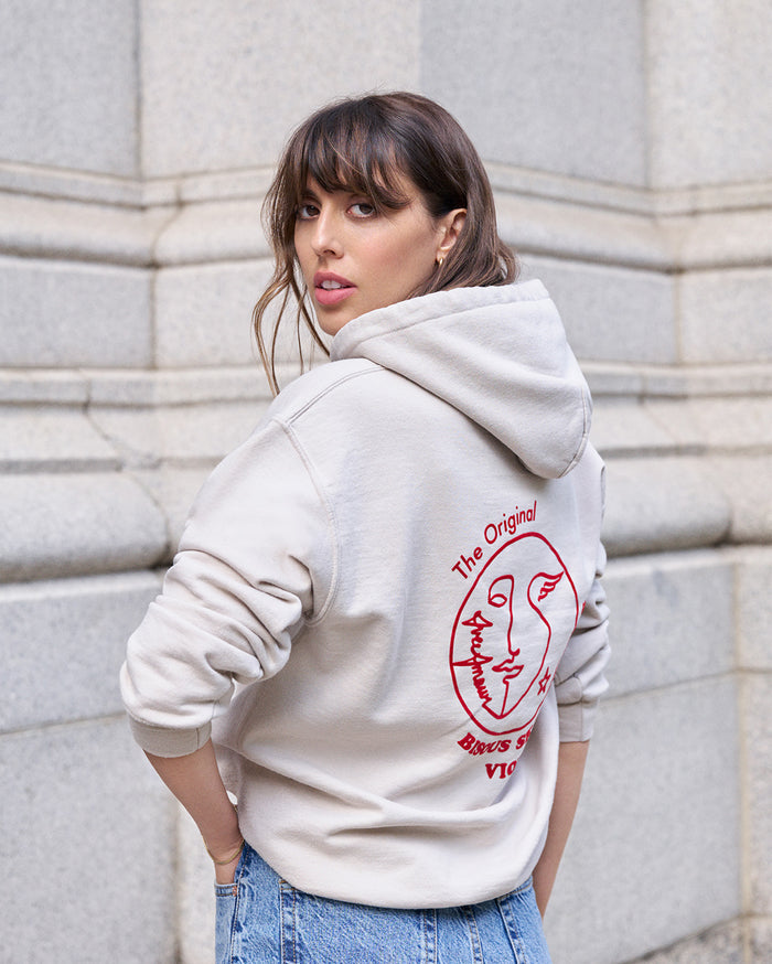 A medium skin tone woman in a light beige hoodie and blue jeans looks over her shoulder. The hoodie's back has a red graphic with an abstract face and the text "The Original BISOUS SANS VIO".