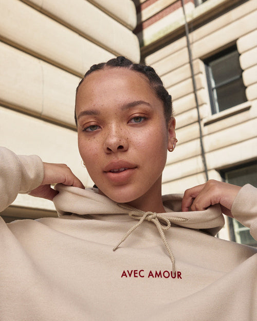 Model with medium skin tone wearing a light beige hoodie with 'AVEC AMOUR' embroidered in red, looking at the camera.