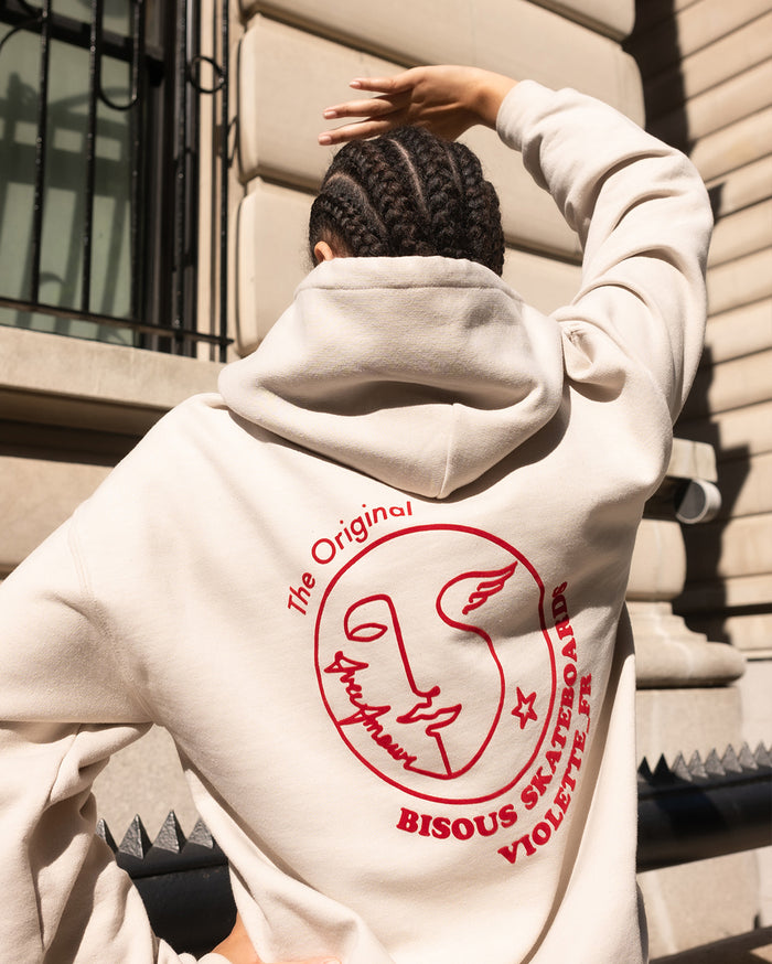 Back of a person with deep skin tone wearing a light beige hoodie. The red graphic on the back reads 'The Original d'Amour SKATEBOARDS VIOLETTE_FR BISOUS' and includes a stylized face and star.