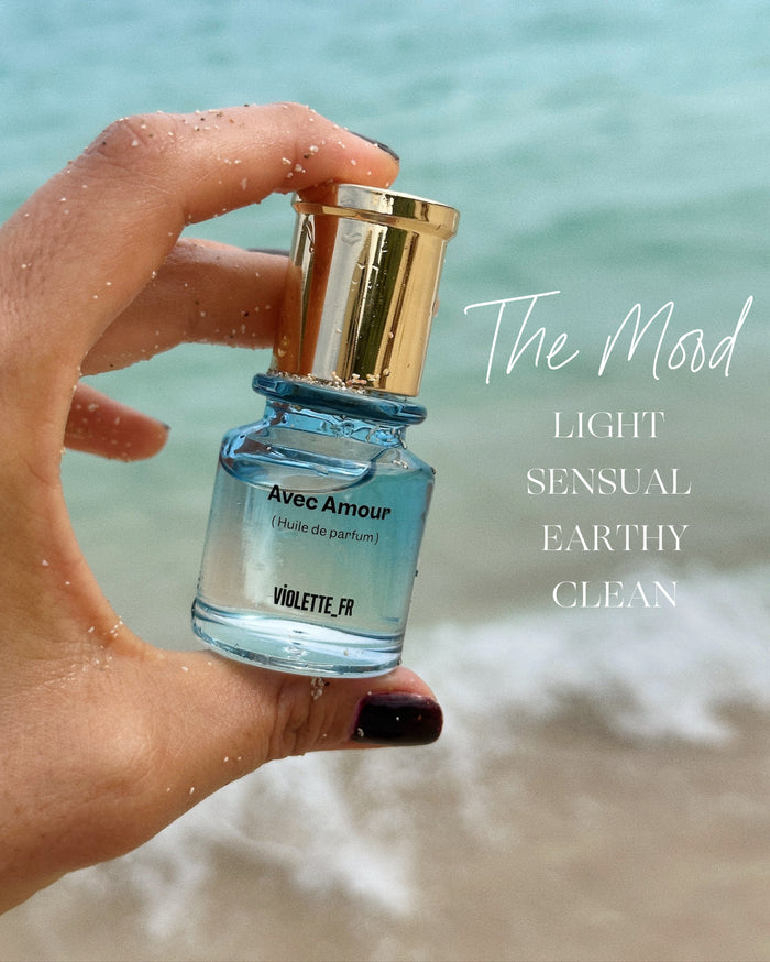 A hand with medium skin tone, covered in sand, holds a small bottle of Avec Amour Huile de parfum by VIOLETTE_FR. The text 'The Mood LIGHT SENSUAL EARTHY CLEAN' is overlaid on a blurred ocean background.