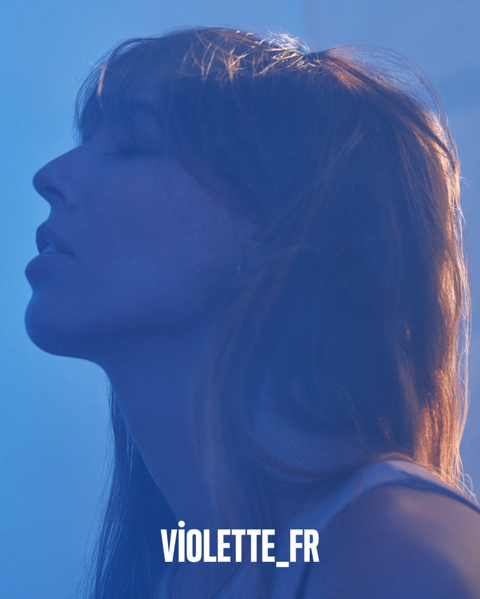 VIOLETTE_FR. Profile of a person with light skin looking upwards, with warm light catching their hair against a blue background.