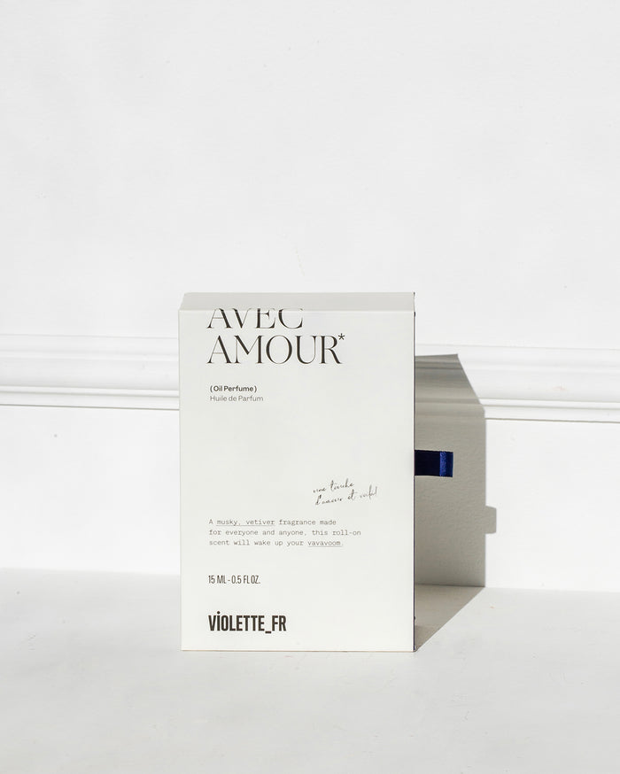 White product box for Violette_FR AVEC AMOUR* (Oil Perfume) Huile de Parfum, 15 ML - 0.5 FL.OZ. Text reads: 'une touche d'amour et voilà!' and 'A musky, vetiver fragrance made for everyone and anyone, this roll-on scent will wake up your vavavoom.' A blue ribbon pull tab is visible.