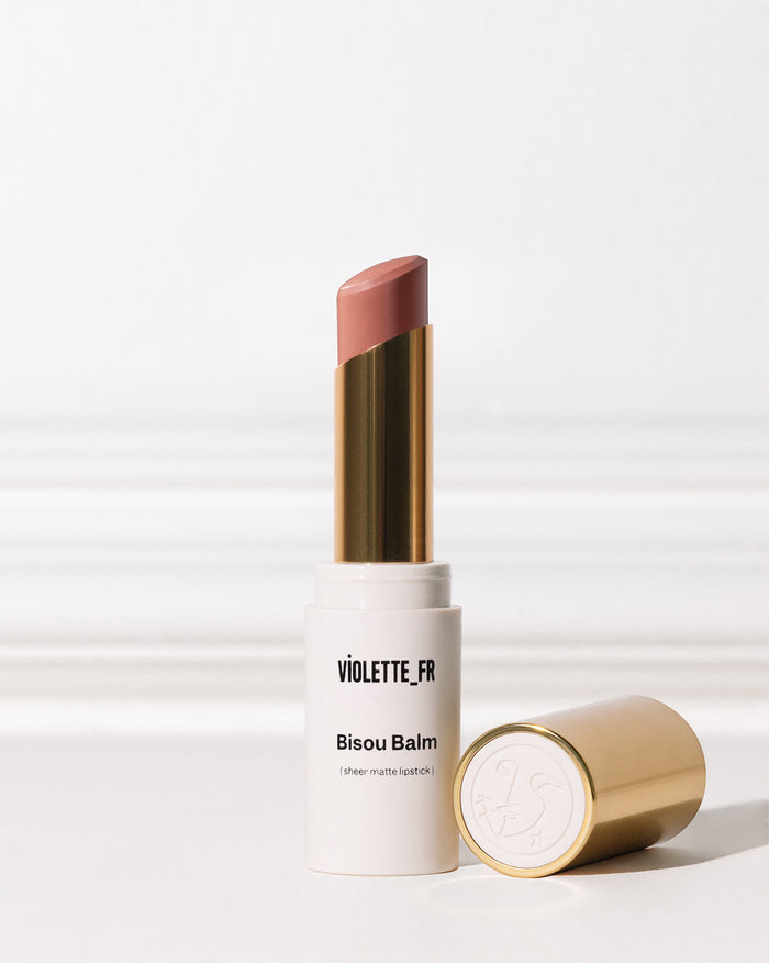 Bisou Balm (sheer matte lipstick) by VIOLETTE_FR, shown in an open white and gold tube with a muted pinkish-brown shade, with its gold cap lying next to it.