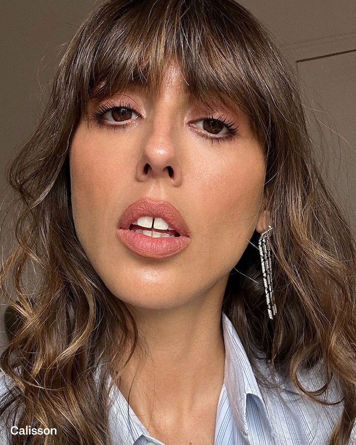 Calisson: Close-up of a woman with medium skin tone, brown wavy hair, and bangs, wearing peachy-nude lipstick and subtle eye makeup, with a long silver earring.
