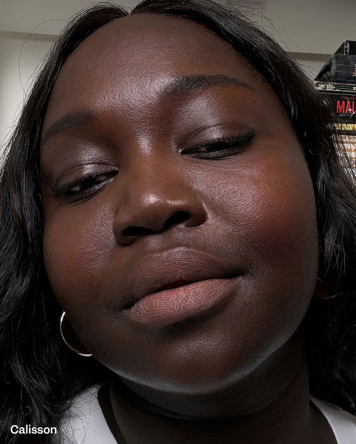 Calisson. Close-up of a model with deep skin tone wearing a subtle, natural-looking lip color and a gold hoop earring.