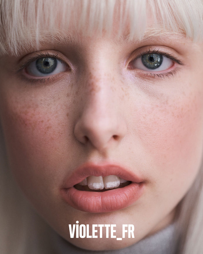 VIOLETTE_FR. Close-up of a model's face with light skin, light blonde bangs, blue-green eyes, and freckles.