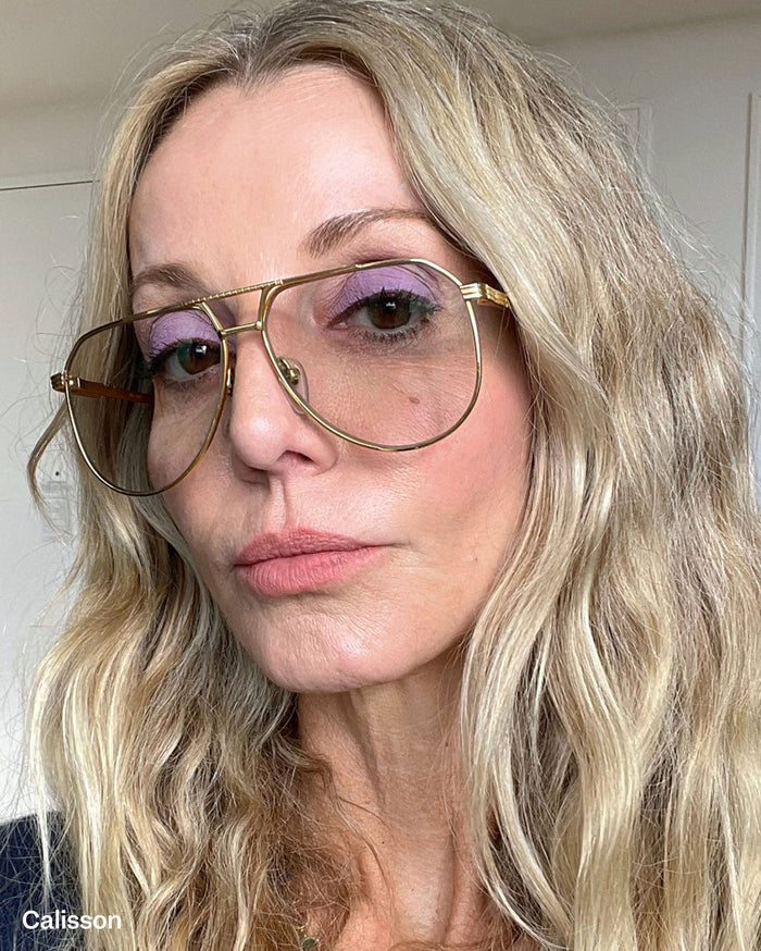 Calisson. Close-up of a woman with light skin tone, blonde wavy hair, wearing large gold-rimmed glasses, purple eyeshadow, and natural pink lipstick.