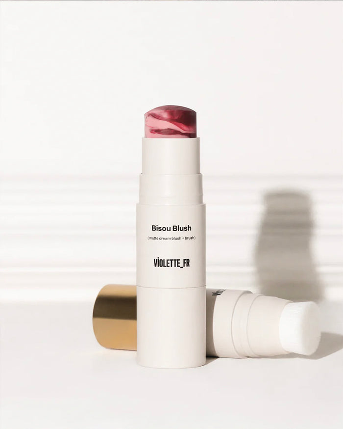 Bisou Blush (matte cream blush + brush) by VIOLETTE_FR. Two light beige blush sticks, one upright showing marbled pink and deep red product, the other horizontal revealing its integrated white brush and gold cap.