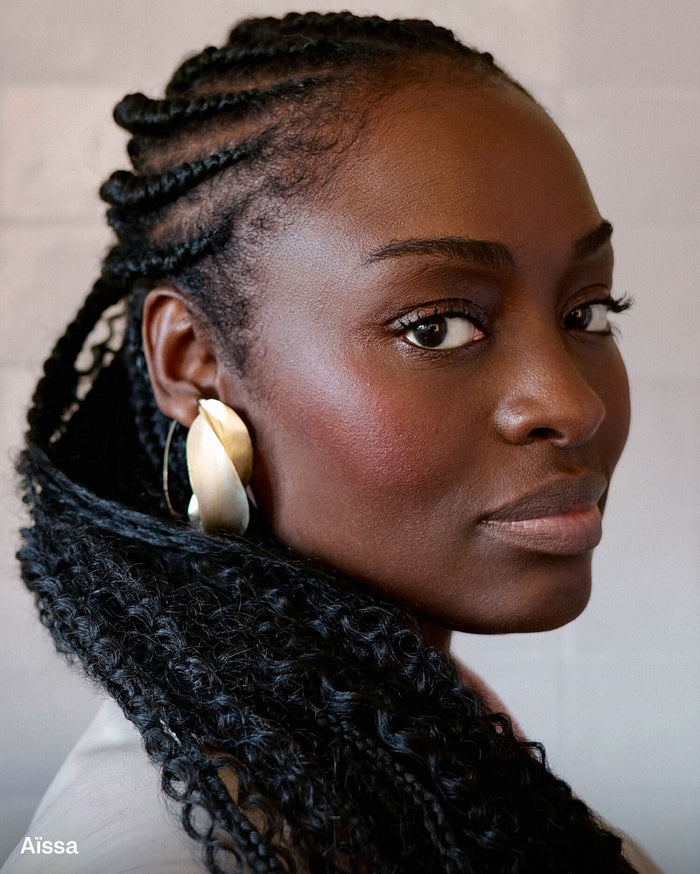 Aïssa, a woman with deep skin tone, wearing a large gold sculptural earring, subtle blush, and natural lip color.