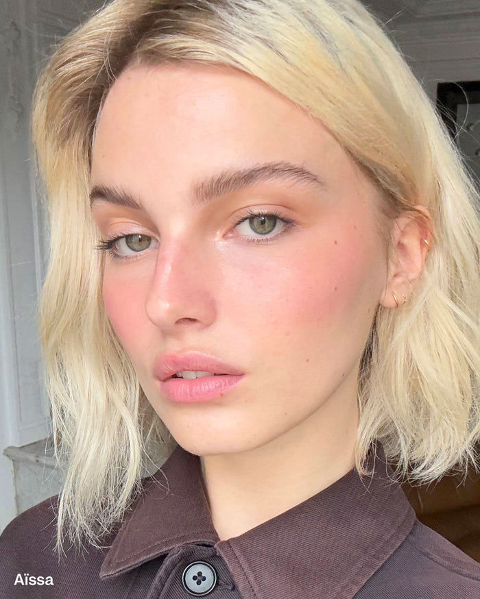 Aissa, a woman with light skin, blonde hair, and green eyes, wears rosy blush and subtle eye makeup with naturally tinted pink lips.