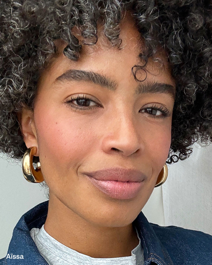 Aïssa, a woman with deep skin tone, wearing soft pink blush, nude-pink lipstick, and large gold hoop earrings.