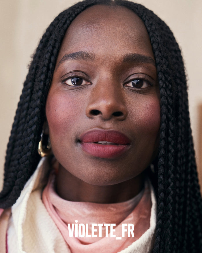 VIOLETTE_FR. Close-up of a woman with deep skin tone, wearing reddish-brown lipstick and blush, with black box braids and a gold hoop earring.