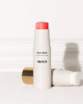Violette_FR Bisou Blush (matte cream blush + brush) sticks. One stands upright, revealing a coral-pink blush; the other lies horizontally, showing a white brush and gold cap.