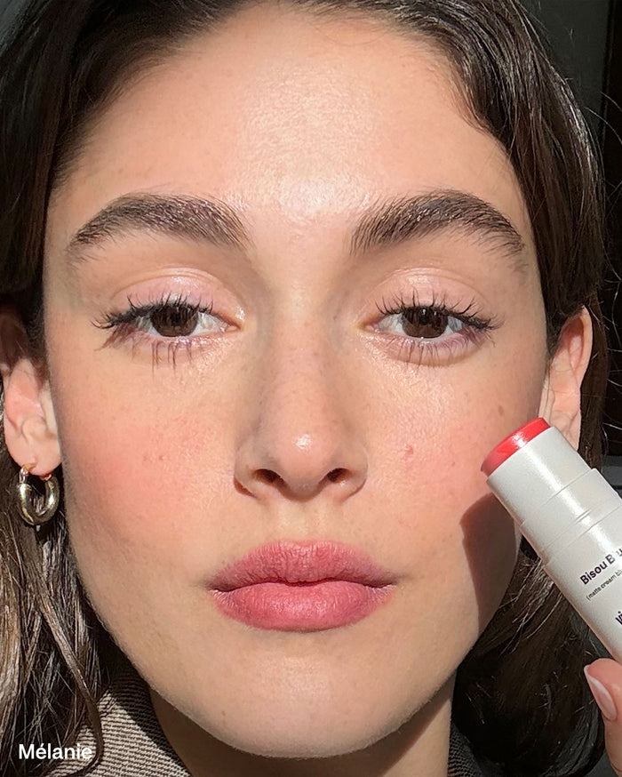 Mélanie, a woman with medium skin tone, wears Bisou Blush (matte cream blush) by violette_fr on her cheeks and lips, holding the coral-red stick to her face.