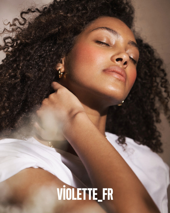 VIOLETTE_FR. Close-up of a woman with medium skin tone, eyes closed, head tilted back, wearing a white t-shirt and gold hoop earrings, with rosy blush and dewy skin.