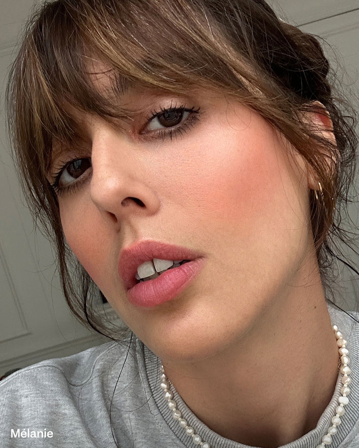 Mélanie. Close-up of a woman with medium skin tone, wearing light pink blush and rosy pink lipstick, a grey top, and a pearl necklace.