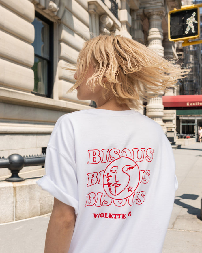 A person with light skin tone wearing a white t-shirt with 'BISOUS BISOUS BISOUS VIOLETTE R' and a circular logo on the back, walking on a city street.