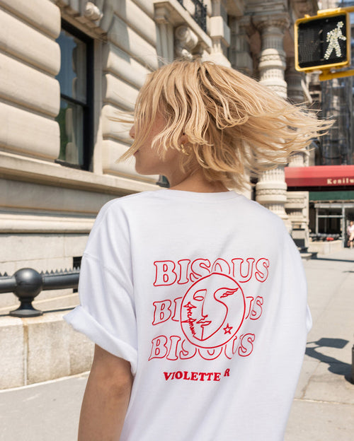 A person with light skin tone wearing a white t-shirt with 'BISOUS BISOUS BISOUS VIOLETTE R' and a circular logo on the back, walking on a city street.