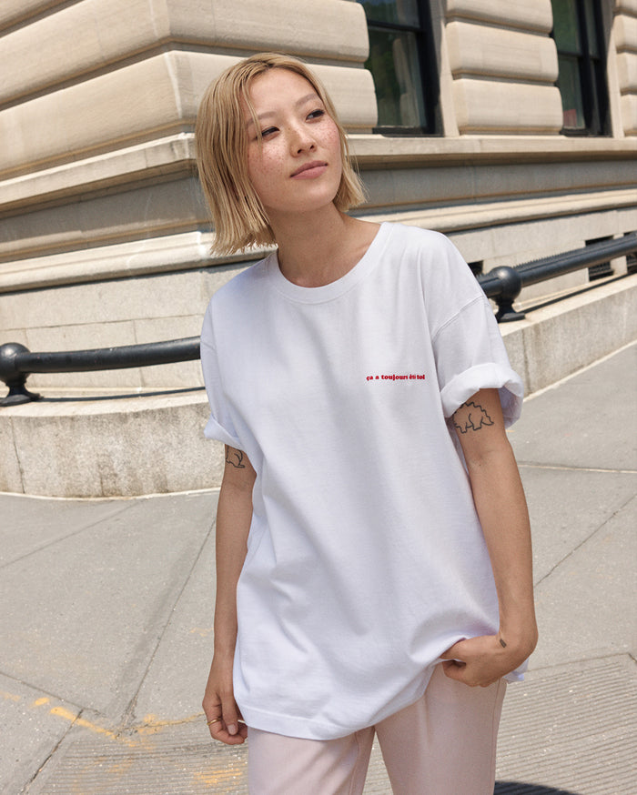 A woman with light skin tone and freckles, with short blonde hair, wears a white t-shirt with red embroidered text 'ça a toujours été toi' on the chest, standing outdoors.