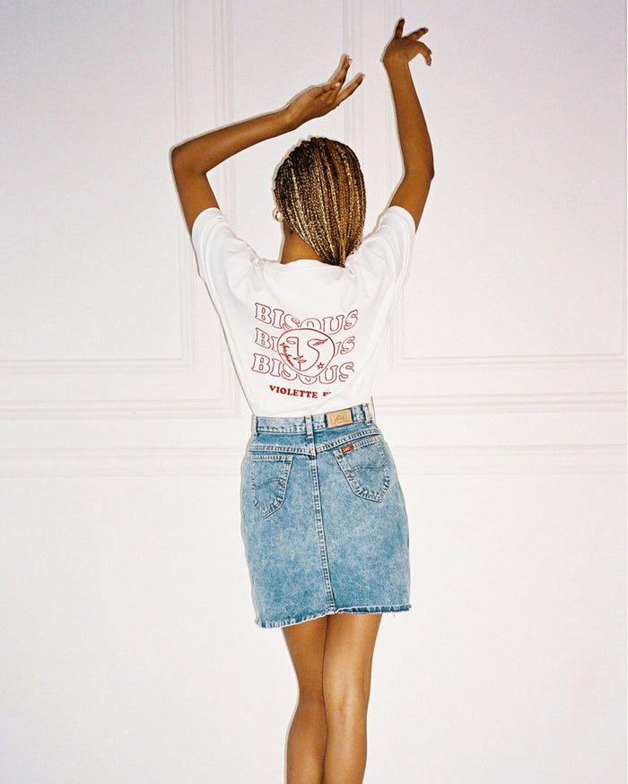 A person with deep skin tone and blonde box braids, seen from the back, wears a white t-shirt with 'BISOUS BISOUS BISOUS VIOLETTE_FR' and a stylized kiss icon, paired with a light blue acid-wash denim mini skirt. Their arms are raised.