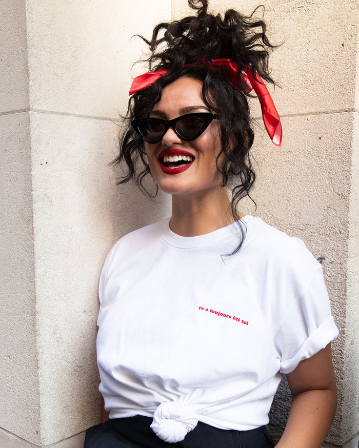 Smiling woman with medium skin tone, red headscarf, sunglasses, and red lipstick, wearing a white t-shirt that reads 'ça a toujours été toi'.