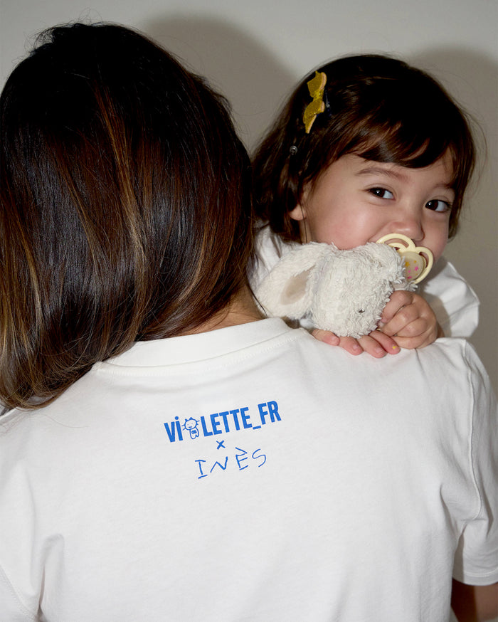 VIOLETTE_FR x INÈS text on the back of a white t-shirt worn by a person with medium skin tone, with a child with light skin tone looking over their shoulder, holding a plush toy and pacifier.