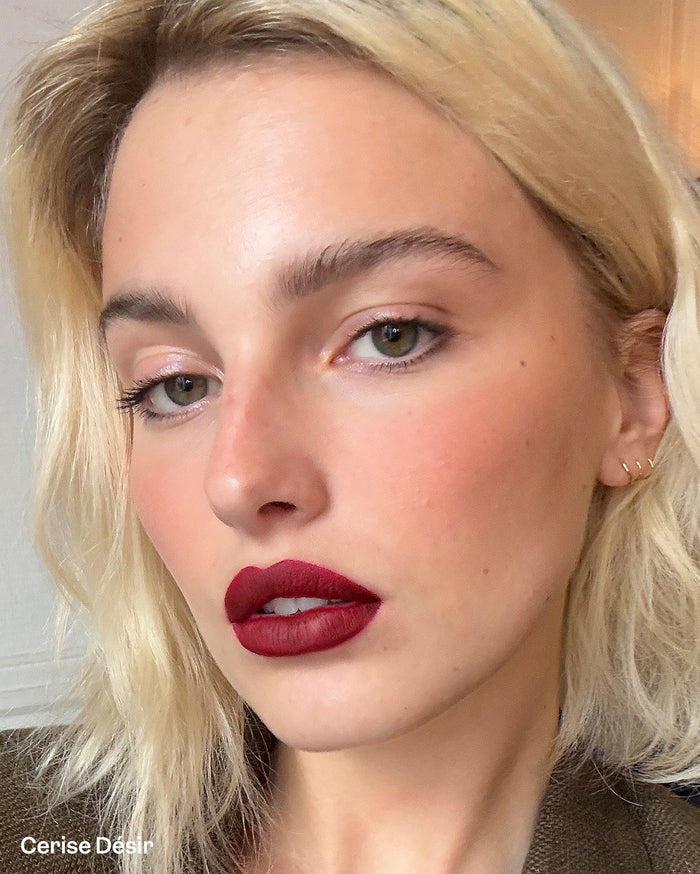 Cerise Désir, a deep red matte lipstick, shown on a woman with light skin, blonde hair, and rosy cheeks.