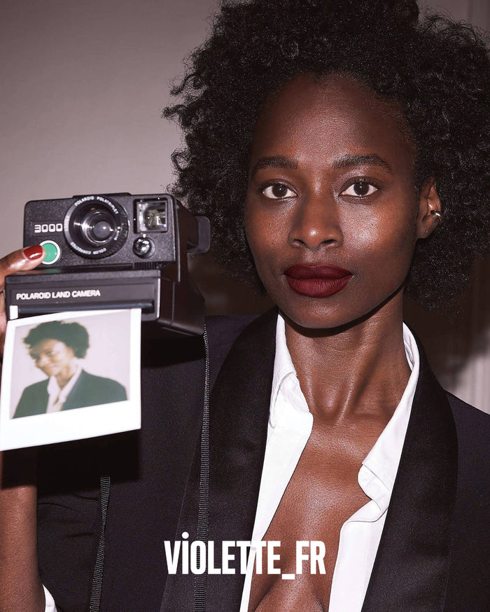 A model with deep skin tone wears dark red lipstick and holds a vintage Polaroid Land Camera 3000, with '3000' and 'POLAROID LAND CAMERA' visible on it. An instant photo is ejecting. The image also features 'VIOLETTE_FR'.