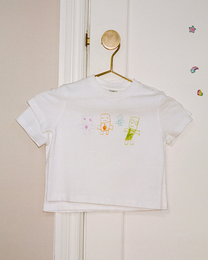 White t-shirt with four colorful, hand-drawn cartoon figures: a pink figure with a purple heart, an orange figure holding a brown cup, a small light blue figure, and a green figure with a green heart. The shirt hangs on a gold hook on a white door with colorful stickers.