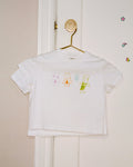 White t-shirt with four colorful, hand-drawn cartoon figures: a pink figure with a purple heart, an orange figure holding a brown cup, a small light blue figure, and a green figure with a green heart. The shirt hangs on a gold hook on a white door with colorful stickers.