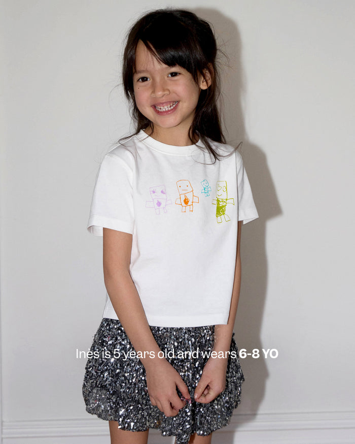 Ines is 5 years old and wears 6-8 YO. A smiling young girl with medium skin tone wears a white t-shirt with colorful drawings and a sparkly silver skirt.