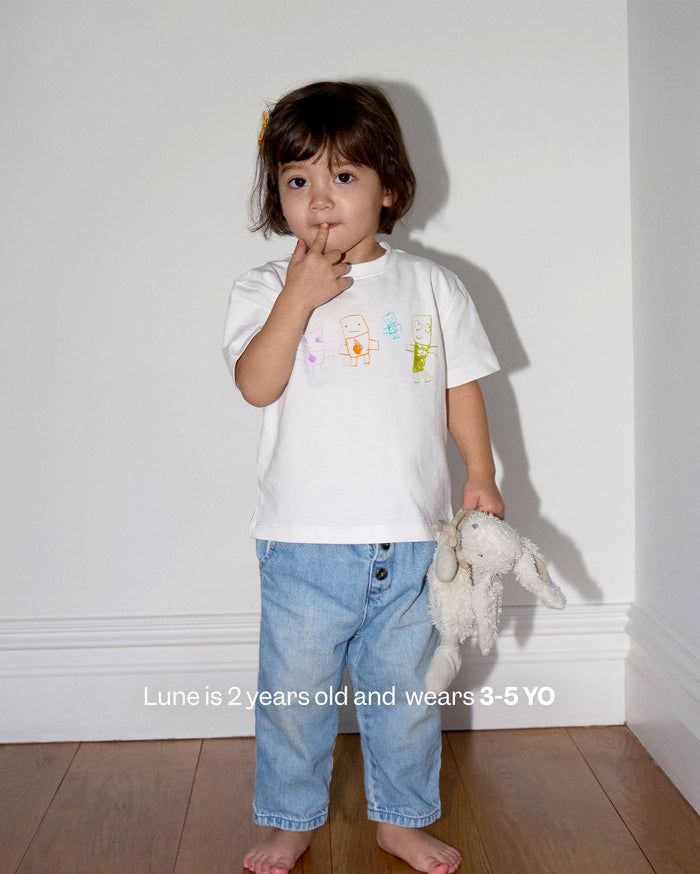 Lune is 2 years old and wears 3-5 YO. A young child with medium skin tone, wearing a white t-shirt with colorful drawings and light blue jeans, holding a plush bunny toy.