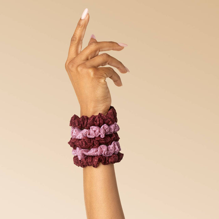 Three fabric scrunchies in burgundy, dusty pink, and mauve are stacked on a person's wrist against a solid beige background.