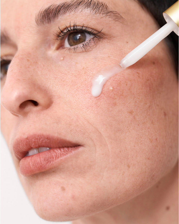 A woman with light to medium skin tone applies a white serum to her cheek with a dropper.