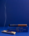 CINNAMON PROJECTS VIOLETTE_FR fragrance on a blue box and cylindrical package of brown incense sticks. A burning incense stick in a gold and blue holder emits smoke against a deep blue background.