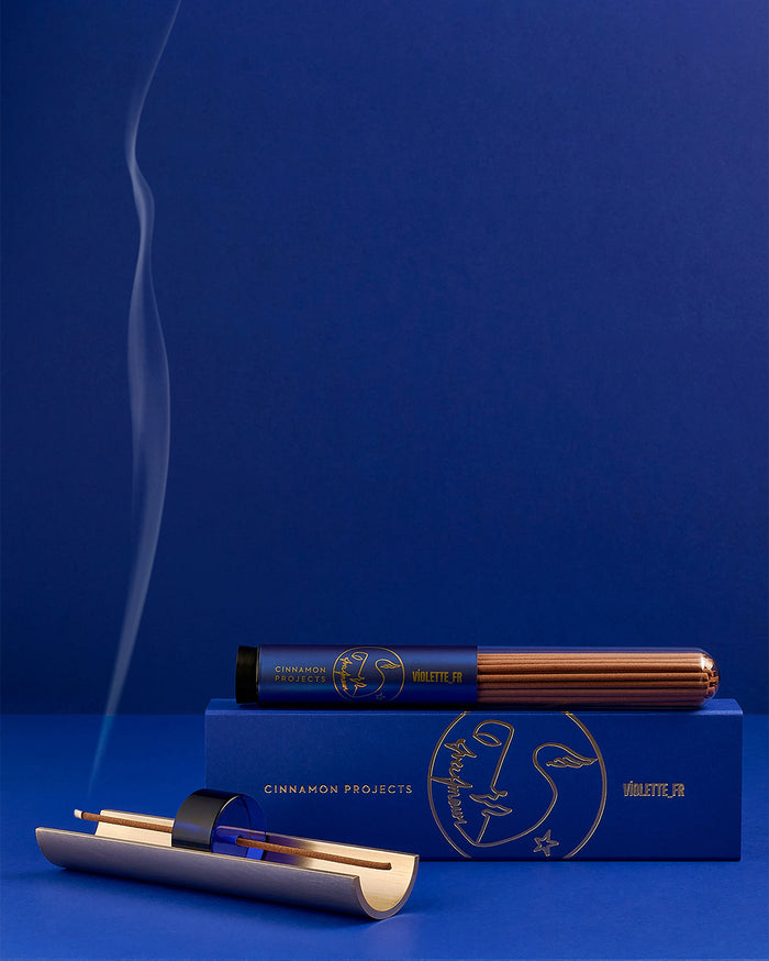 CINNAMON PROJECTS VIOLETTE_FR fragrance on a blue box and cylindrical package of brown incense sticks. A burning incense stick in a gold and blue holder emits smoke against a deep blue background.