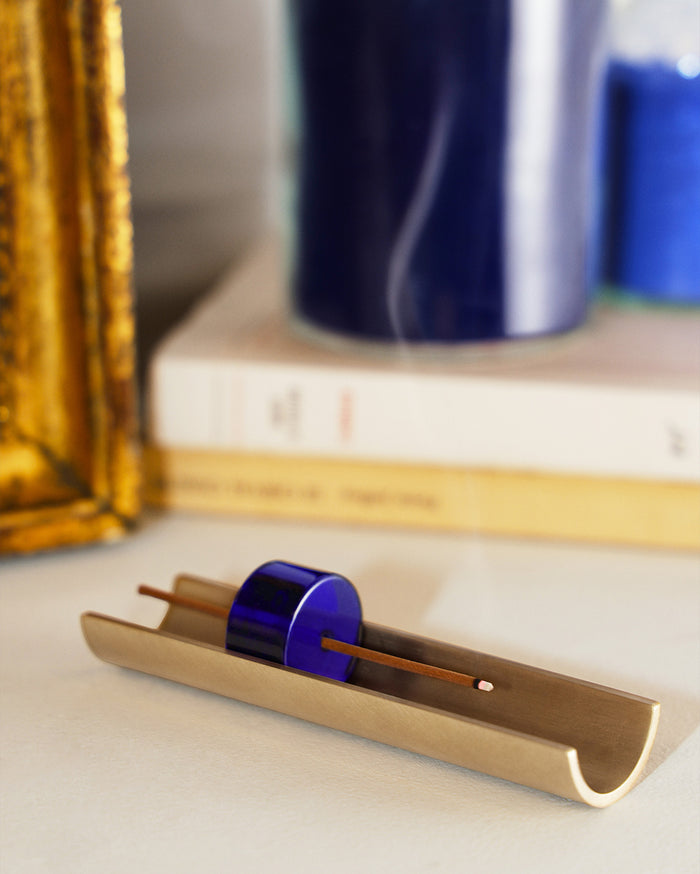 A golden-bronze trough-shaped incense holder with a dark blue glass bead securing a burning brown incense stick.