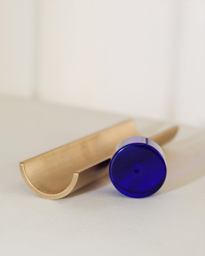 A deep blue cylindrical jar lies on its side next to a golden-bronze curved metallic object, all resting on a light off-white surface.
