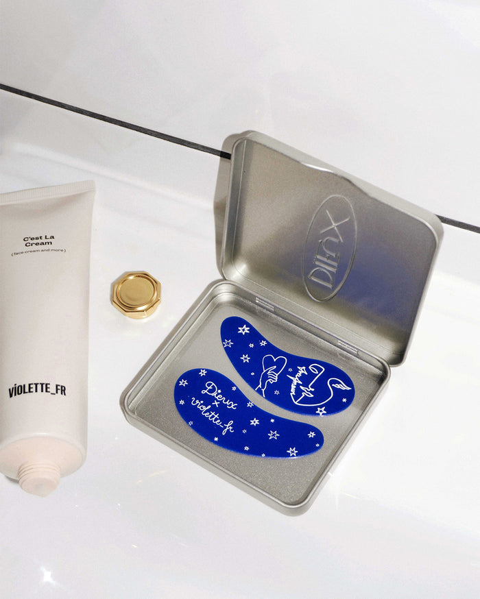 Violette_FR C'est La Cream tube and a gold cap next to an open silver Dieux tin containing two blue eye patches. The patches feature white designs of stars, a hand holding a heart, a face, a swan-like figure, and the text 'Dieux violette.fr' and 'Faire l'Amour'.