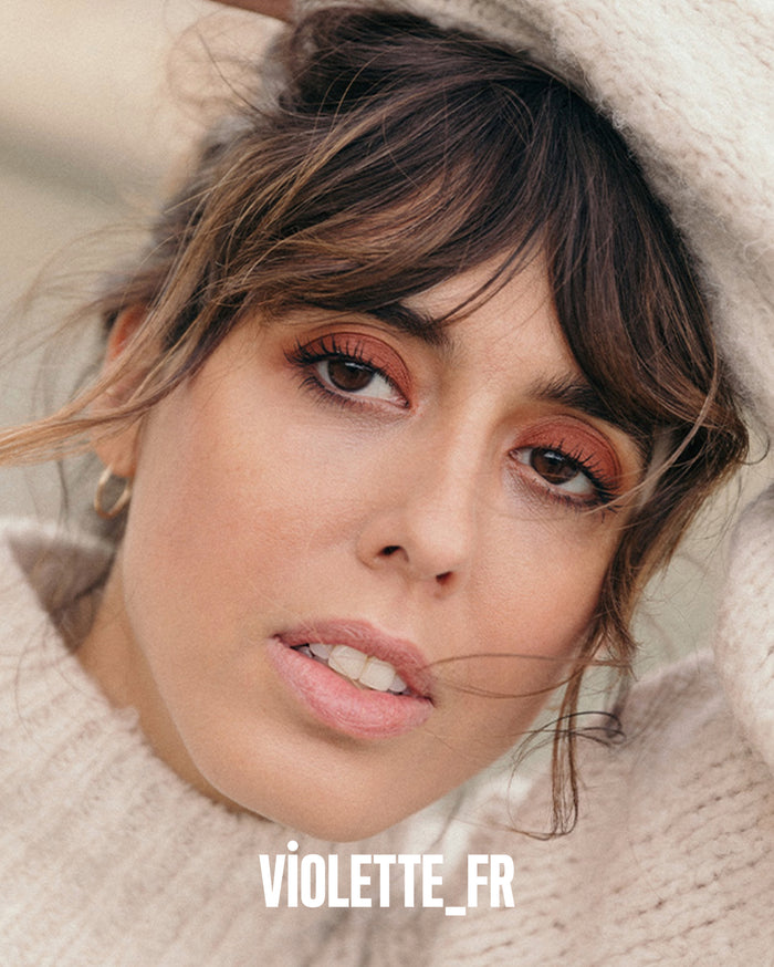 VIOLETTE_FR. Close-up of a woman with medium skin tone, wearing a cream knitted sweater and warm reddish-brown eyeshadow.