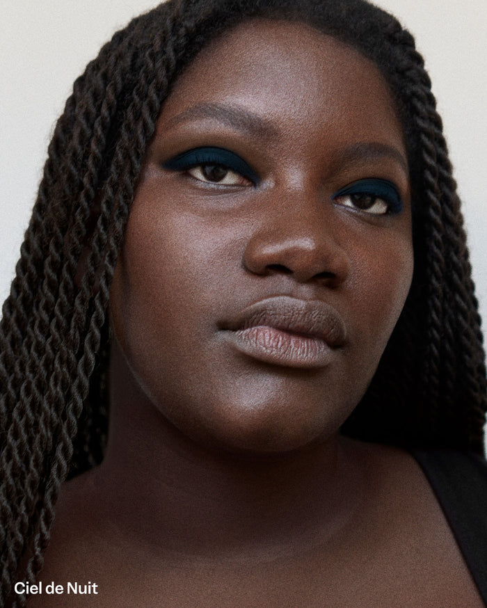 Ciel de Nuit: Close-up portrait of a person with deep skin tone and braided hair, wearing a deep teal eyeliner on their upper and lower lash lines.