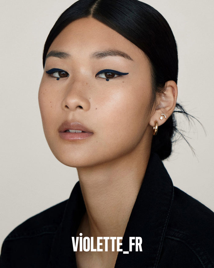 VIOLETTE_FR. A model with medium skin tone wears dark blue winged eyeliner with a dot under each eye, a black denim jacket, and gold earrings.