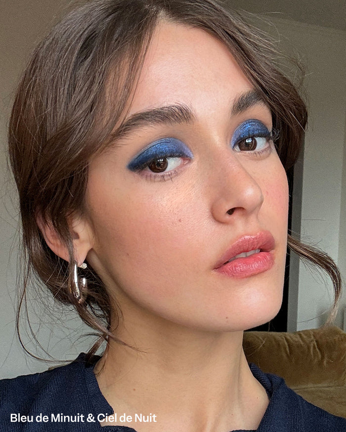 Bleu de Minuit & Ciel de Nuit. Close-up of a person with medium skin tone wearing vibrant, shimmery blue eyeshadow and glossy natural pinkish-nude lipstick.