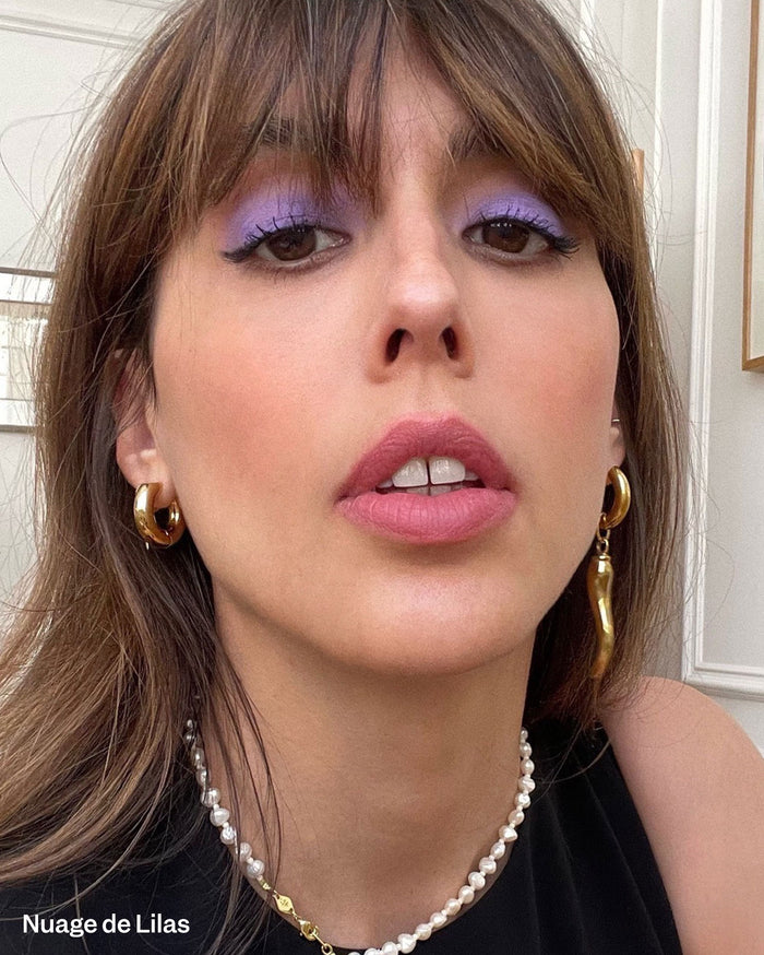 Nuage de Lilas. A woman with medium skin tone wears lilac eyeshadow, black eyeliner, and matte pink lipstick, accessorized with gold earrings and a pearl necklace.