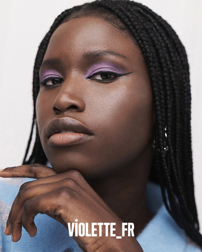 VIOLETTE_FR. Close-up of a deep skin tone model with braided hair, wearing vibrant purple eyeshadow and black winged eyeliner, with her hand resting under her chin.