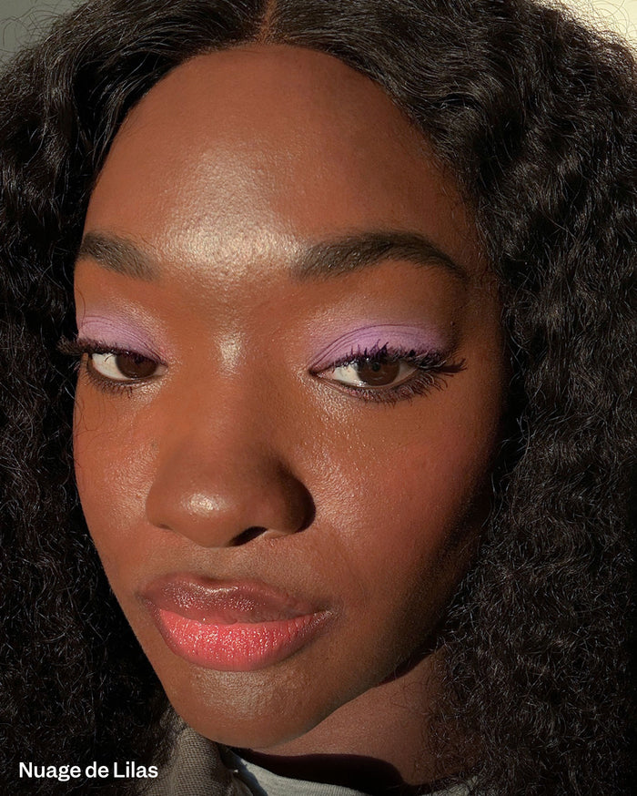 Nuage de Lilas. Close-up of a model with deep skin tone, wearing light purple eyeshadow and peachy-pink lip gloss.