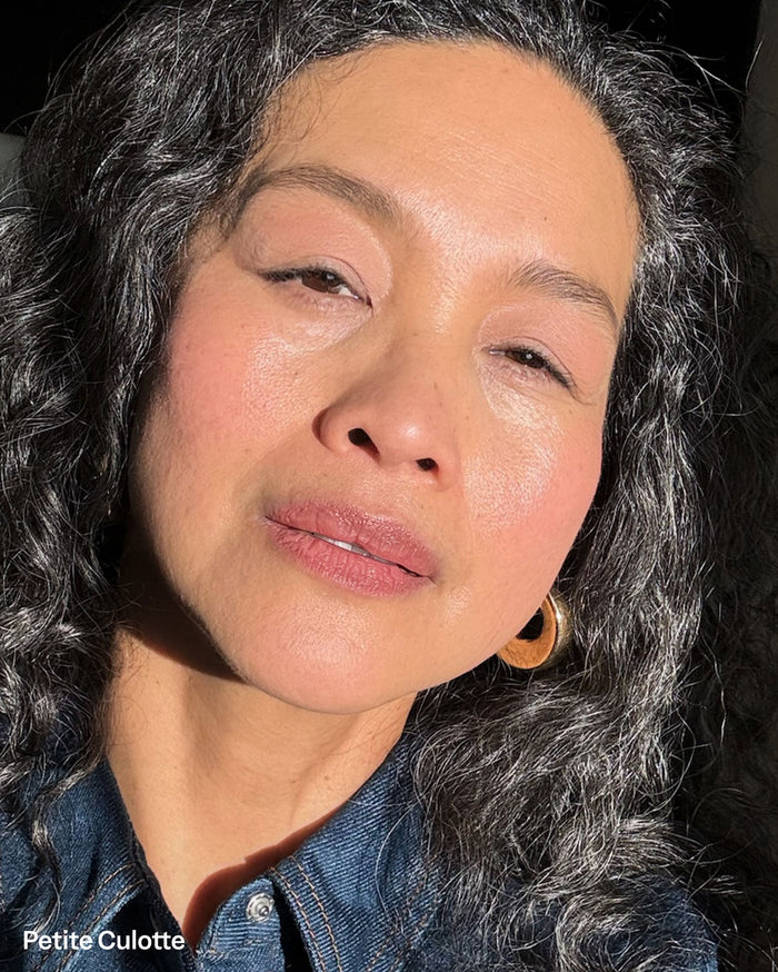 Petite Culotte. Close-up of a woman with medium skin tone, showcasing a soft pink blush on her cheeks and a matching lip color. She has dark curly hair with grey strands, wears a denim shirt, and a gold hoop earring. Sunlight illuminates her face.