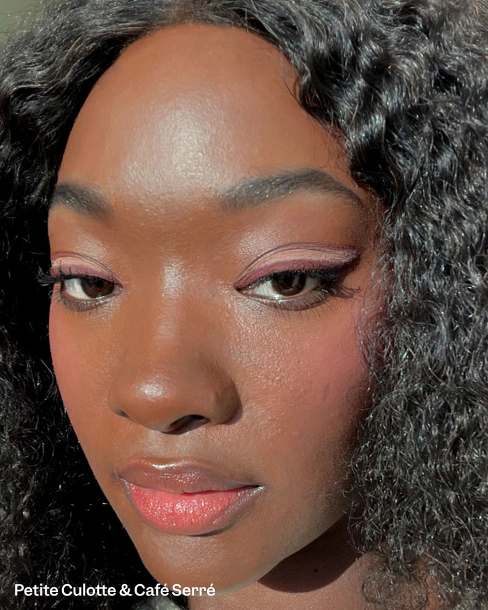 Petite Culotte & Café Serré. Close-up of a person with deep skin tone, wearing reddish-plum winged eyeliner and peachy-pink glossy lipstick.